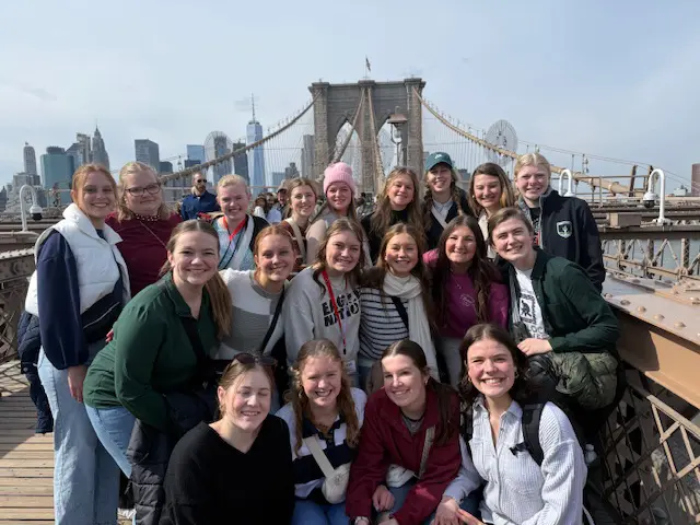 Students posing on the Brooklyn Bridge with the Manhattan skyline in the background.